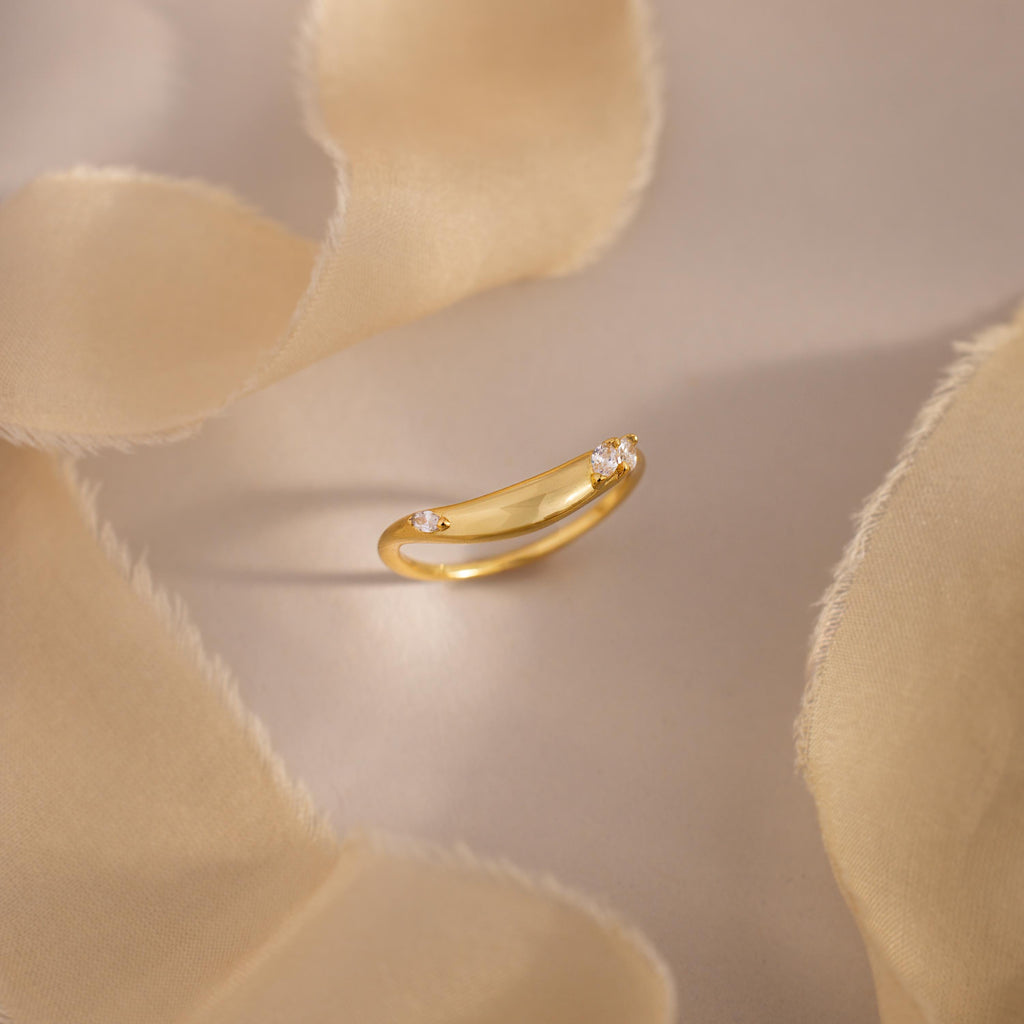 Gold curved band ring with three marquise cut clear gemstones, displayed on soft beige fabric for an elegant and minimal jewelry presentation.