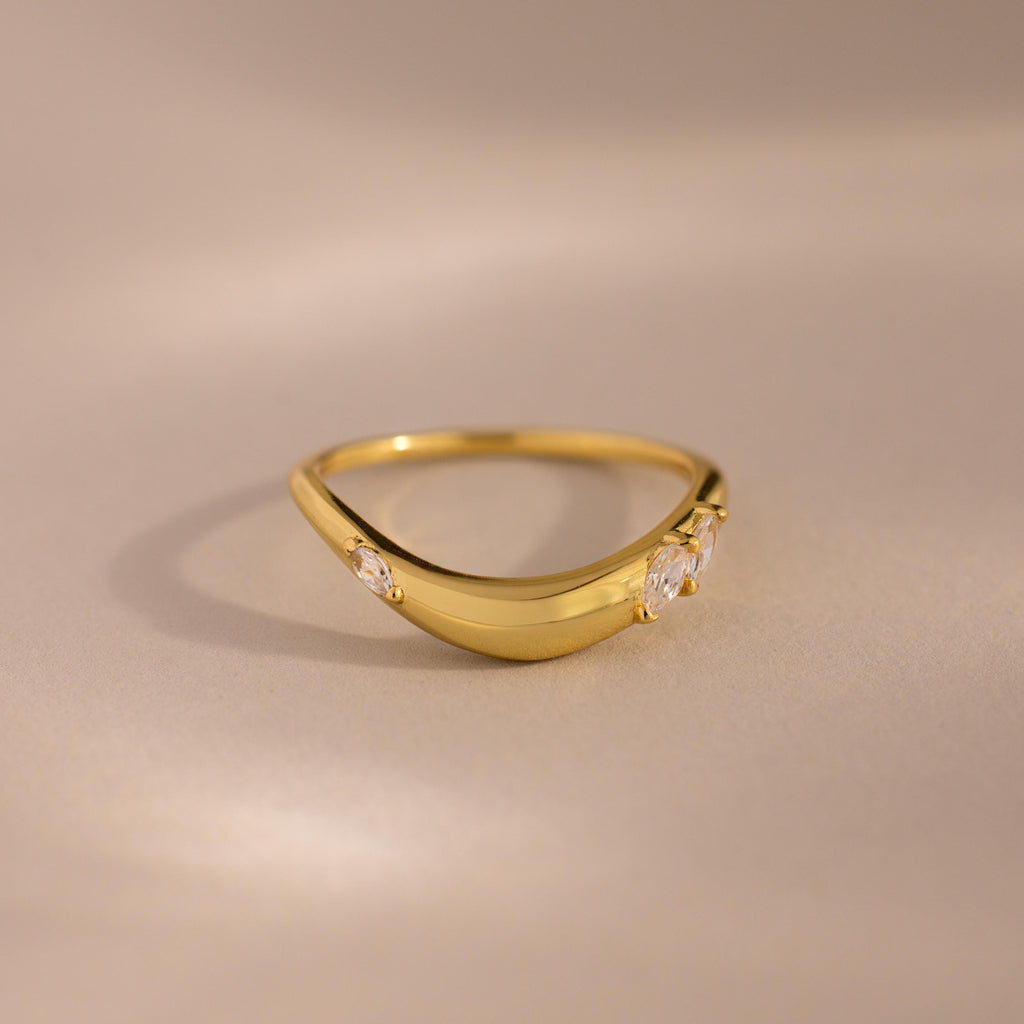 Gold curved band ring with three marquise cut clear gemstones set along the front, displayed on a neutral beige background.