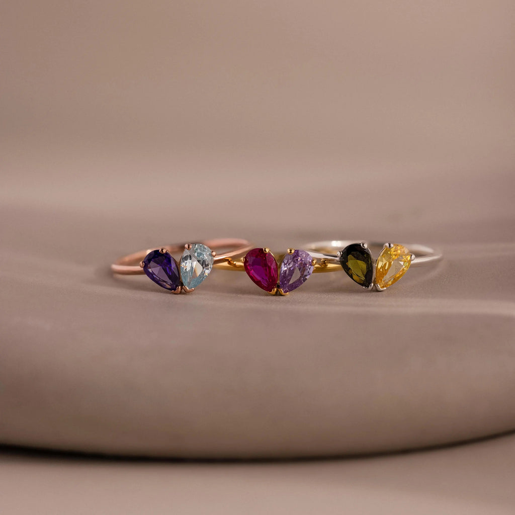 The Toi et Moi Teardrop Heart Ring in Rose Gold features three delicate heart rings with pear-cut stones in purple, blue, pink, black, and yellow, shimmering gracefully on a neutral background.