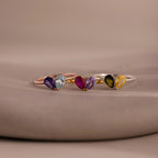 The Toi et Moi Teardrop Heart Ring in Rose Gold features three delicate heart rings with pear-cut stones in purple, blue, pink, black, and yellow, shimmering gracefully on a neutral background.