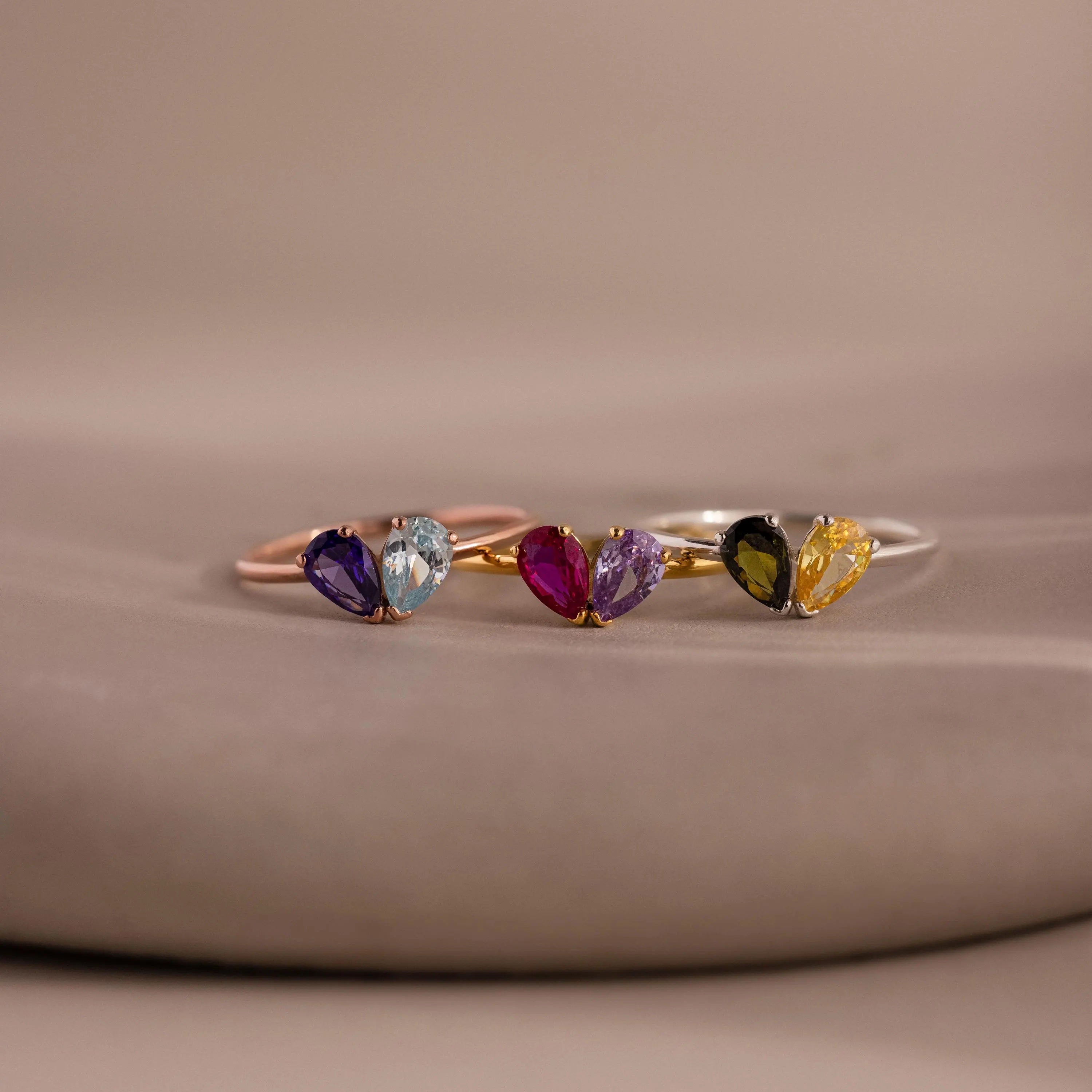 The Toi et Moi Teardrop Heart Ring in Rose Gold features three delicate heart rings with pear-cut stones in purple, blue, pink, black, and yellow, shimmering gracefully on a neutral background.