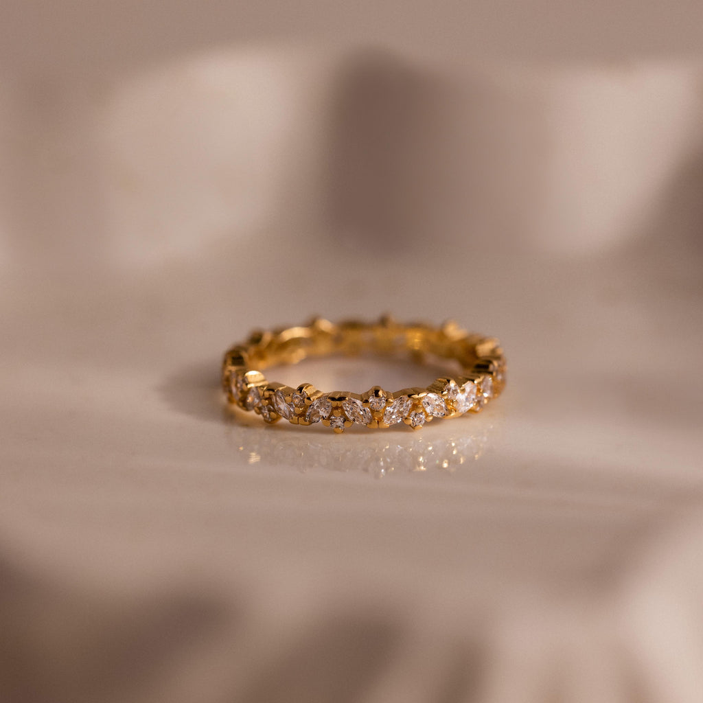 The Diamond Cluster Eternity Ring, featuring sparkling diamonds encircling the band, is displayed on a soft, cream-colored surface.