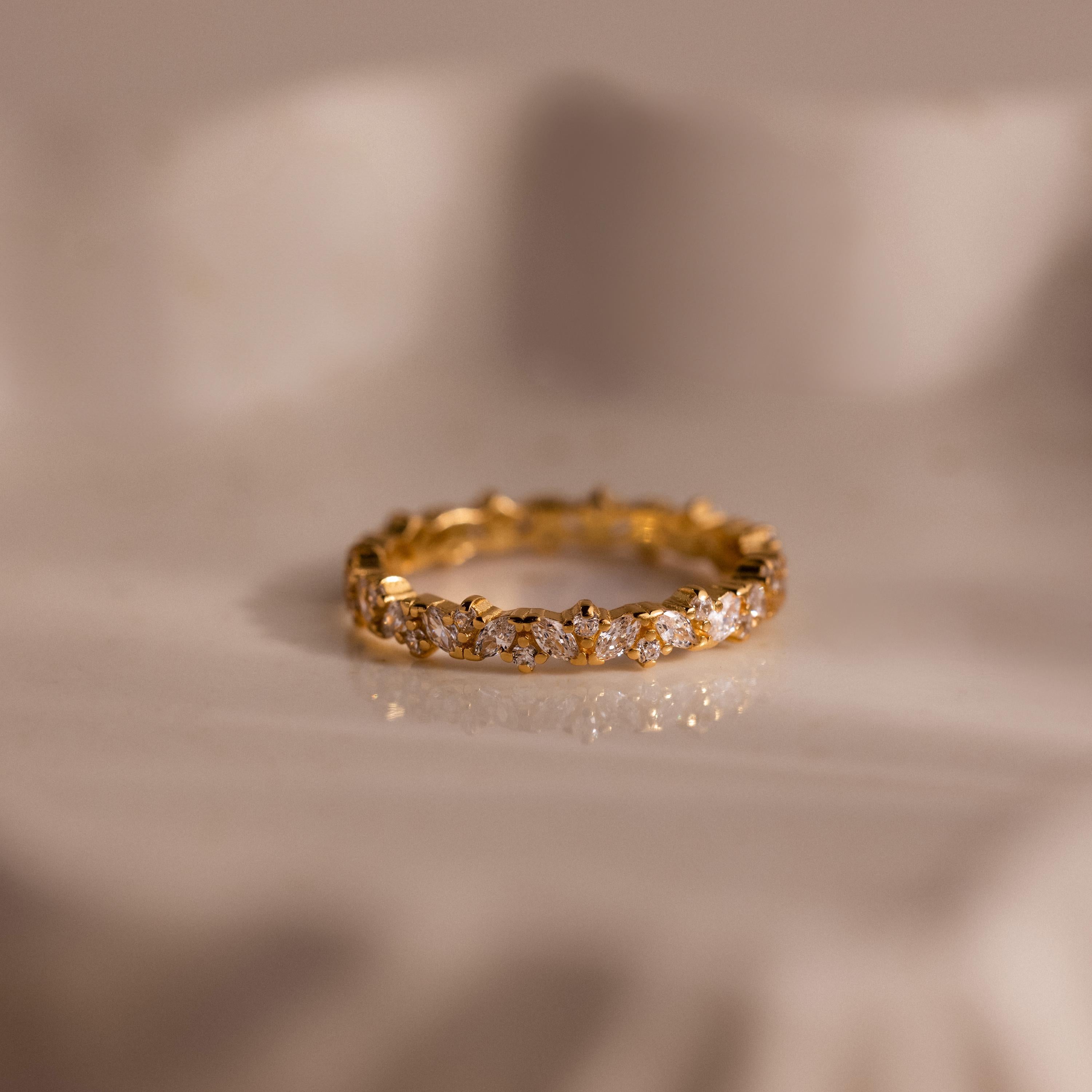 The Diamond Cluster Eternity Ring, featuring sparkling diamonds encircling the band, is displayed on a soft, cream-colored surface.