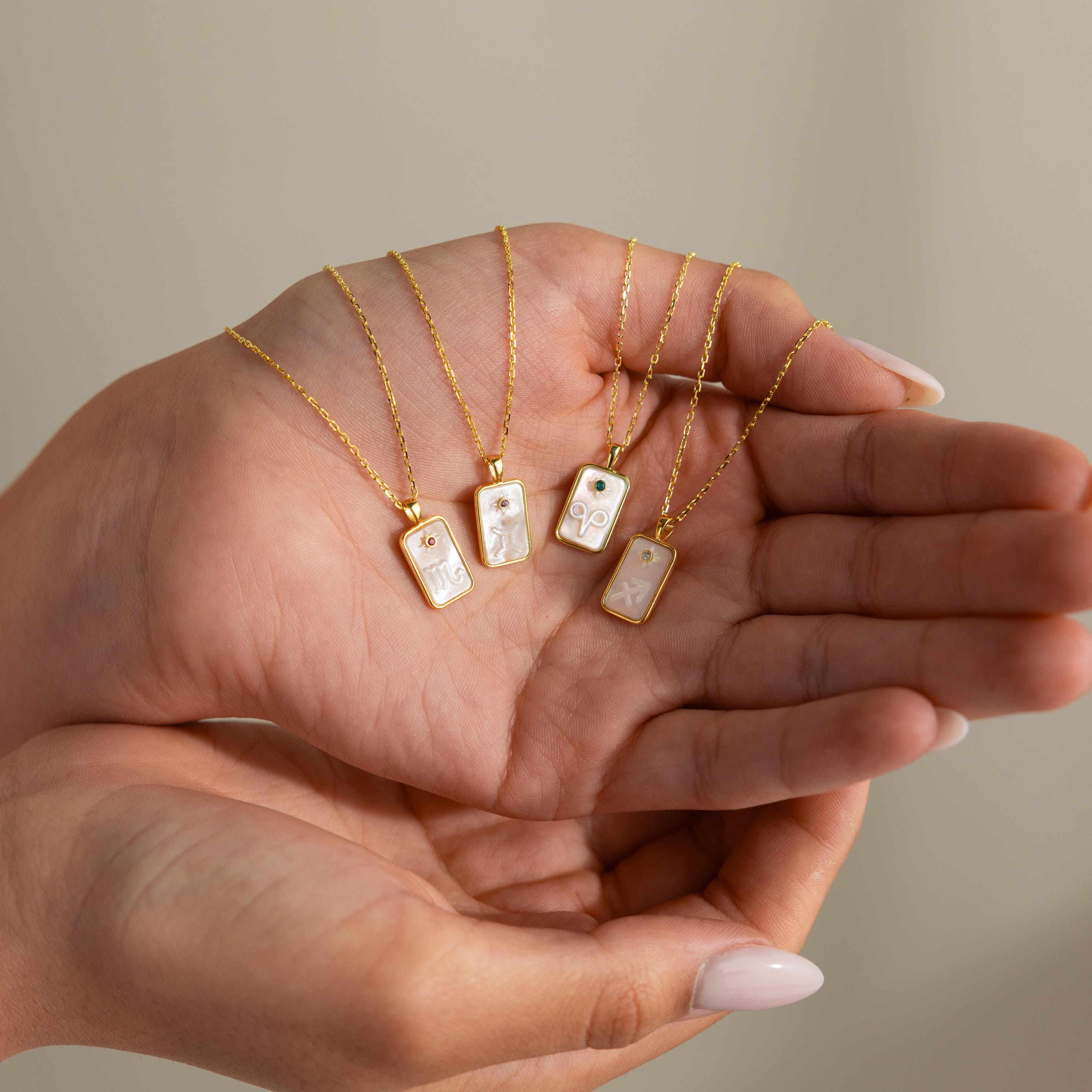 A hand holds four Pearl Zodiac Birthstone Necklaces in 18K Gold, each featuring rectangular pendants with zodiac symbols—ideal as personalized jewelry or a unique birthstone necklace.