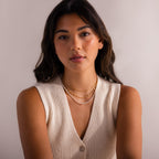 Woman wearing layered gold chain necklace with delicate white pearl beaded strands, styled with a cream knit sleeveless top for an elegant minimal look.