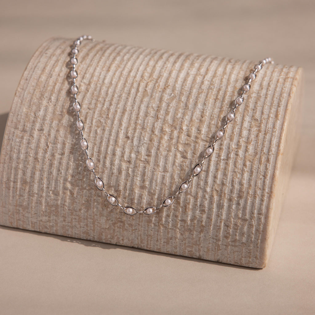 Silver pearl chain necklace with white pearls set in oval silver links, displayed on textured stand for an elegant, modern presentation.