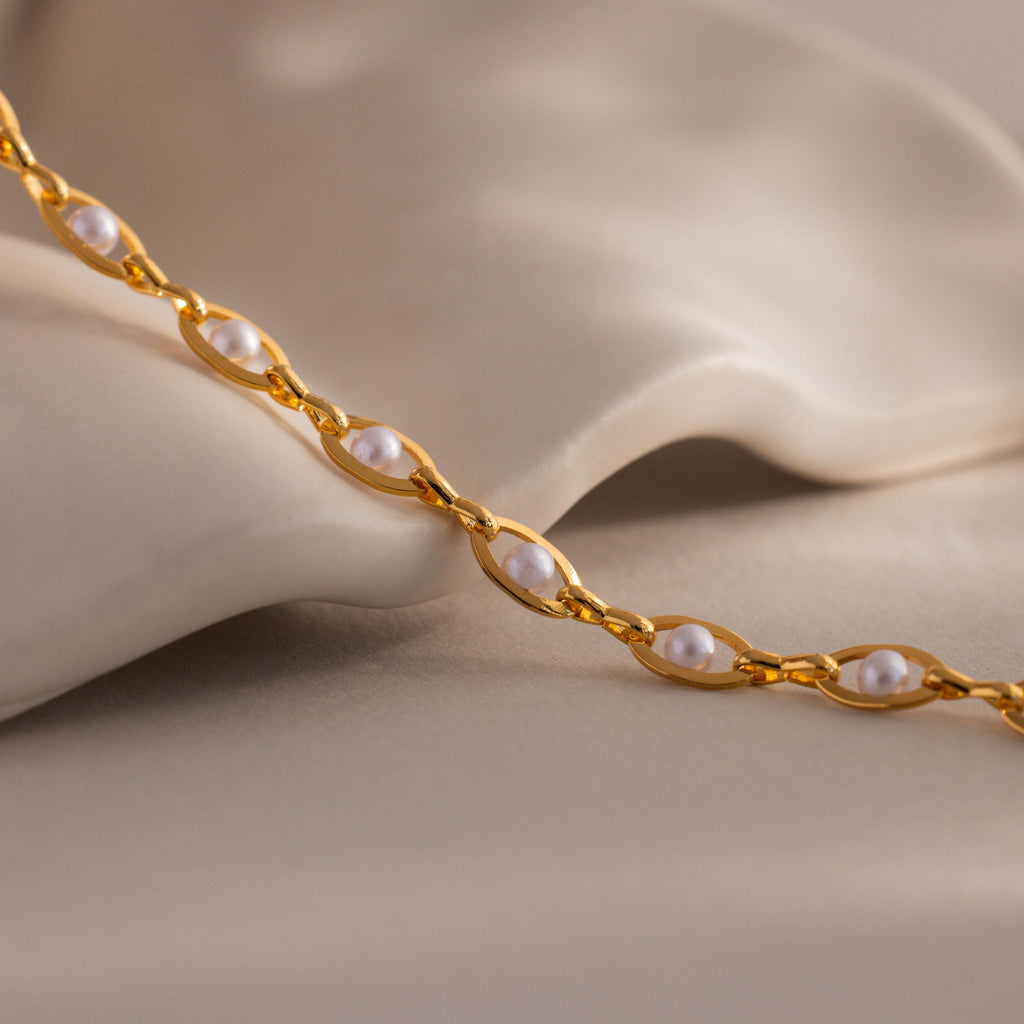 Close up of a gold pearl chain necklace with small white pearls set in oval gold links, displayed on a soft neutral fabric background.