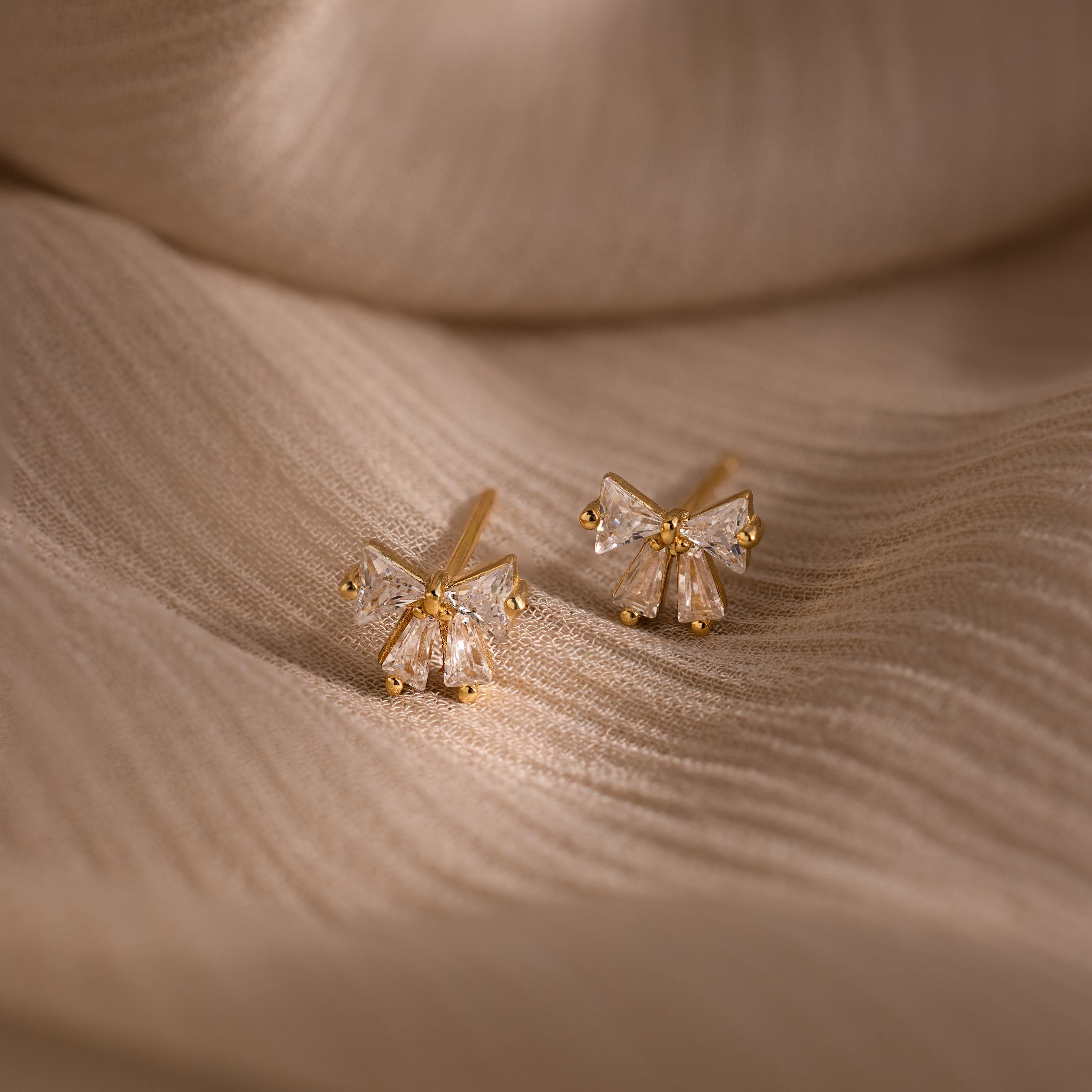 Gold Starlight Bow stud earrings with baguette cut clear stones in a delicate bow design, displayed on a soft beige fabric for an elegant and refined presentation.