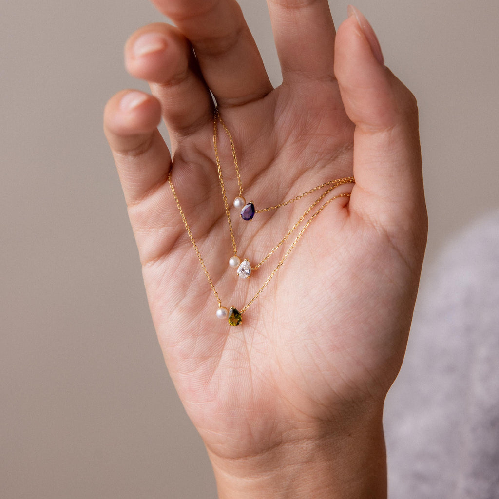 A hand holds three delicate gold necklaces, including the Teardrop Garnet & Pearl Necklace featuring purple, green, and white gemstone pendants—an ideal milestone gift.
