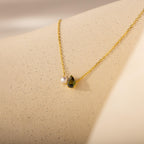 A timeless gift, the Teardrop Peridot & Pearl Necklace features a pearl and green gemstone pendant, beautifully displayed on a light, textured surface.