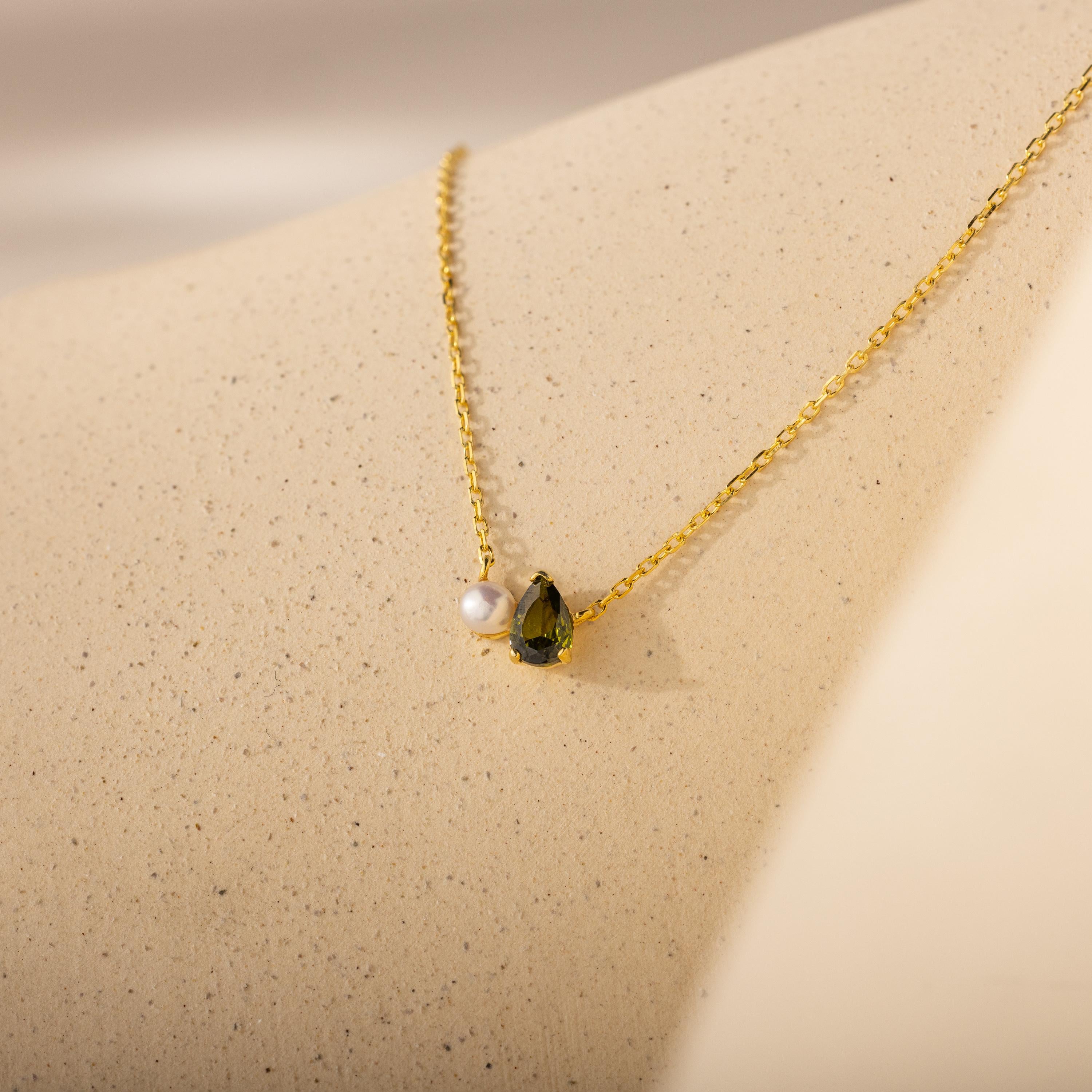 A timeless gift, the Teardrop Peridot & Pearl Necklace features a pearl and green gemstone pendant, beautifully displayed on a light, textured surface.