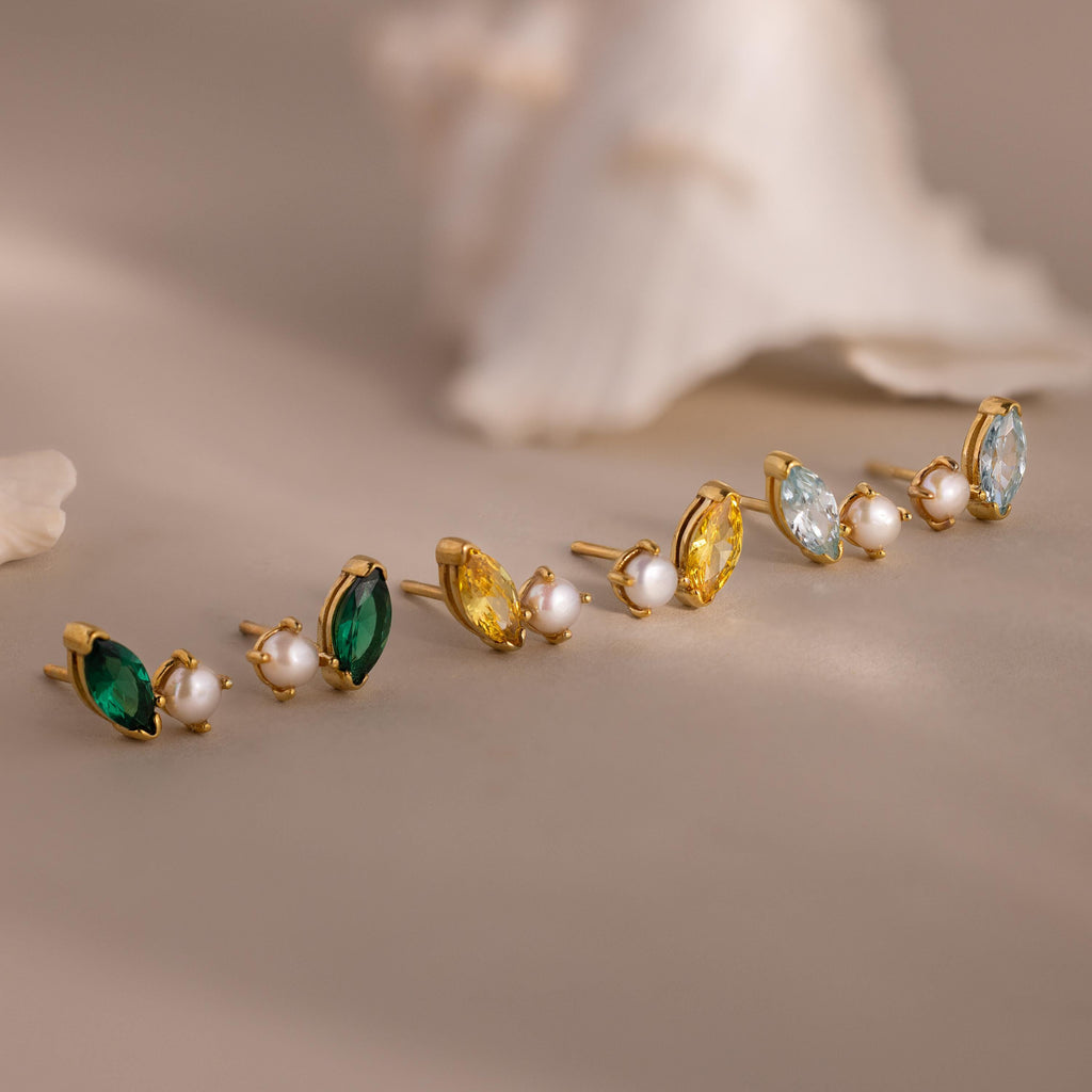 The Marquise Birthstone & Pearl Studs set features three pairs of gold stud earrings with marquise birthstones in green, yellow, and blue accents, plus classic pearl studs, elegantly arranged on a beige surface.