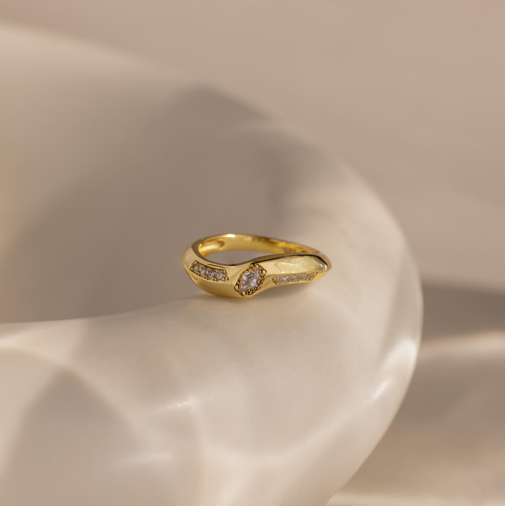 Radiant Facet Ring in polished gold with an diamond shaped CZ stone center and rows of pave set side stones, displayed on a smooth ivory surface.