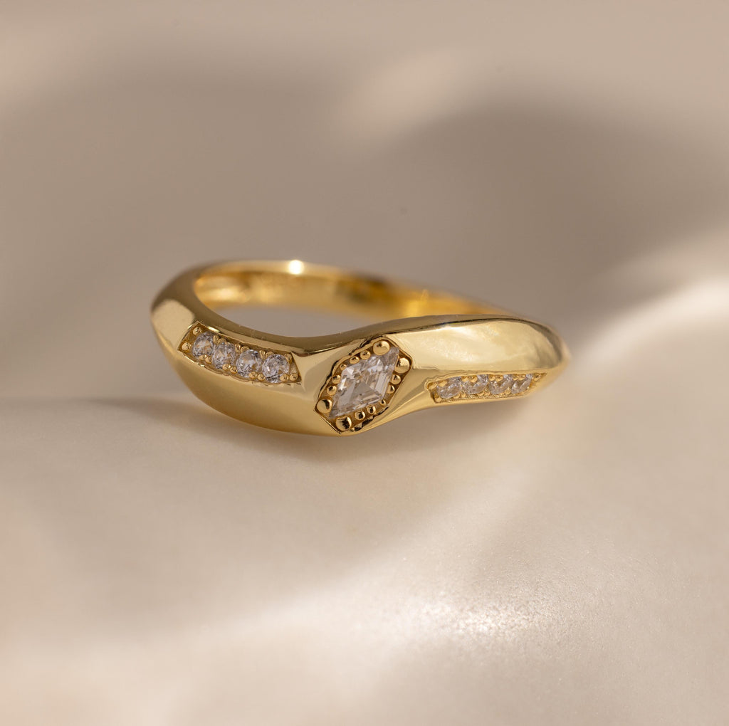 Close up of the Radiant Facet Ring in polished gold featuring a diamond shaped CZ stone in center with pave set side stones, displayed on a smooth ivory surface.