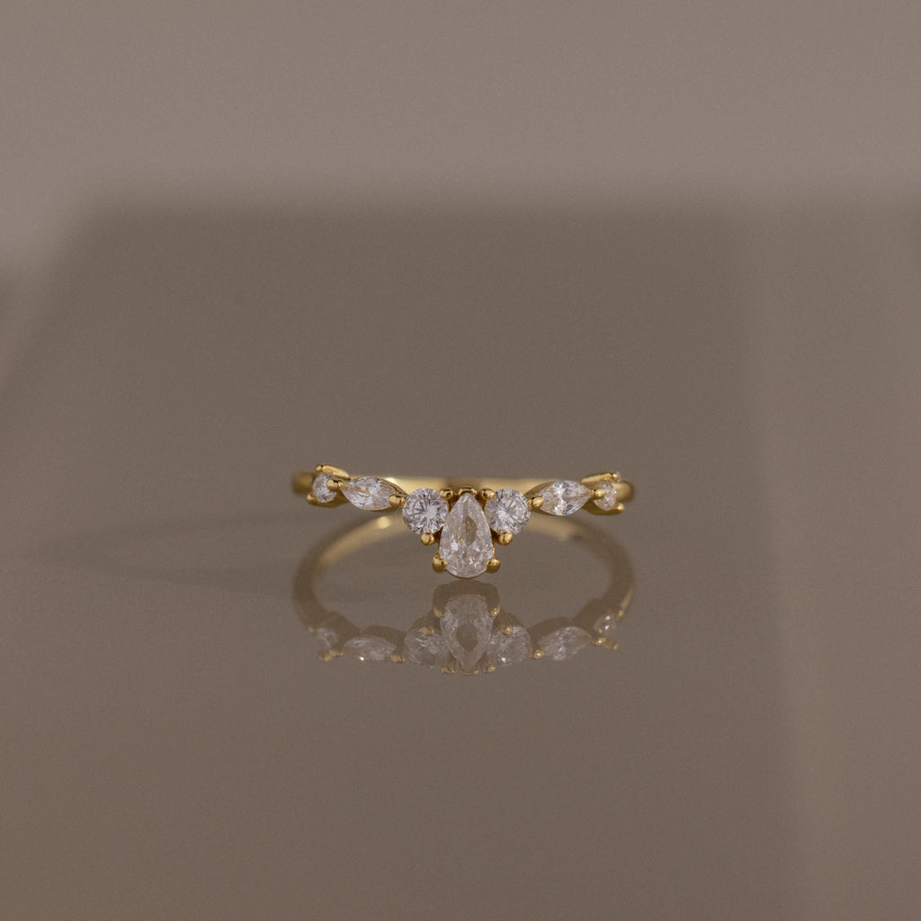Radiant Tear Ring in polished gold featuring a pear cut diamond center accented by round and marquise diamonds, displayed on a reflective neutral surface.