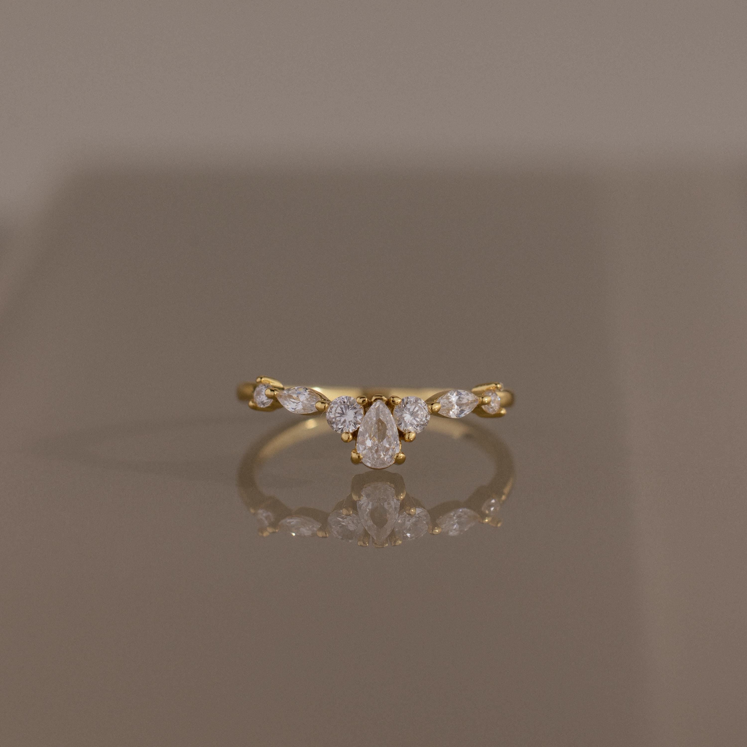 Radiant Tear Ring in polished gold featuring a pear cut diamond center accented by round and marquise diamonds, displayed on a reflective neutral surface.