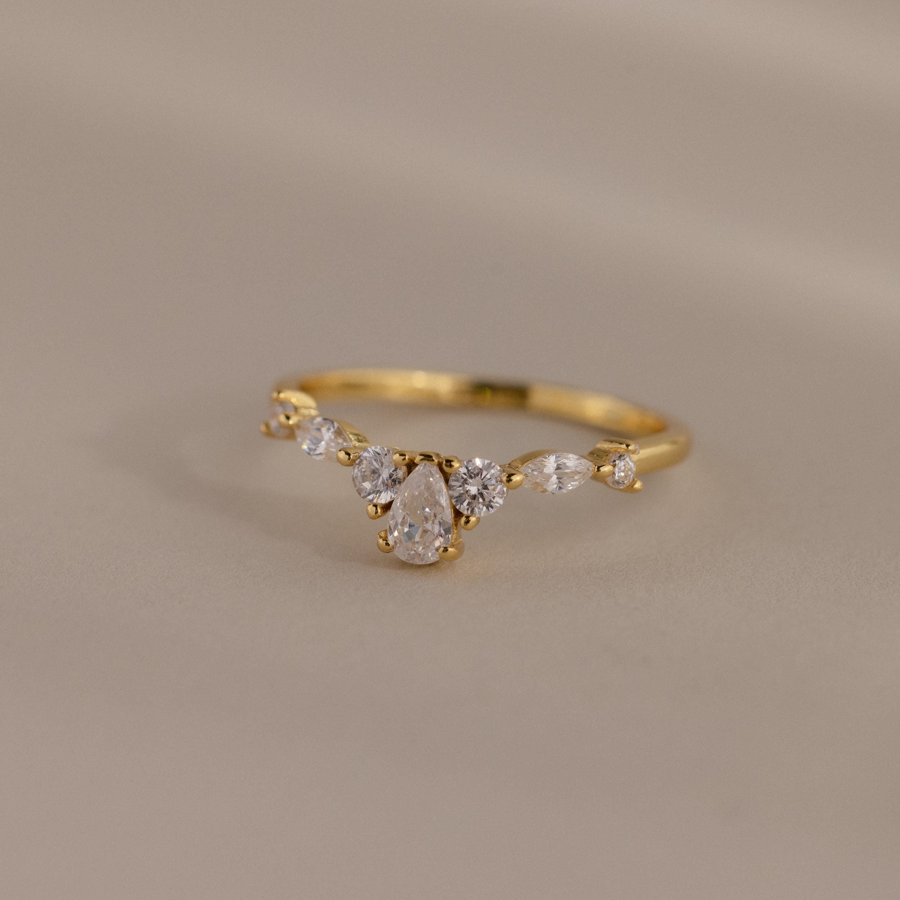 Radiant Tear Ring in polished gold with a pear cut diamond center framed by round and marquise side stones, displayed on a soft neutral background.