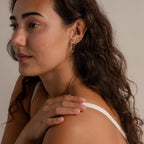 Woman with long curly hair wearing a white strap top and vintage-inspired Bow Chain Studs gold earrings, touching her shoulder.
