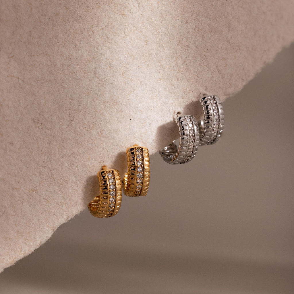 A pair of Ribbed Pave Huggies hoop earrings in gold and silver displayed on a light, textured surface—ideal for statement jewelry lovers.