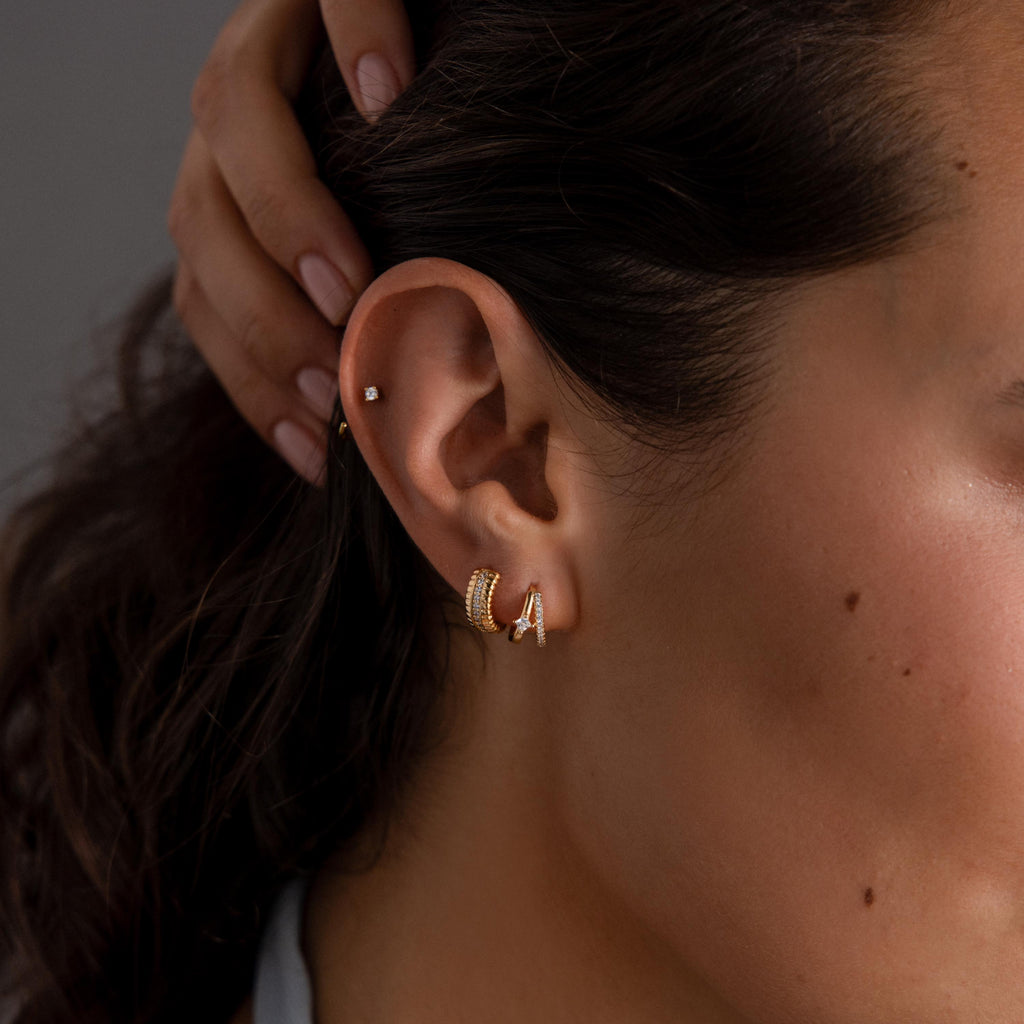 Close-up of a womans ear wearing Ribbed Pave Huggies in 18K Gold, featuring chunky hoops and a small stud on the upper ear, showcasing elegant gold tones.