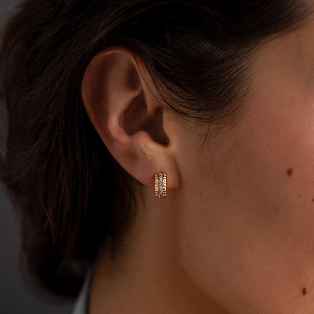 Close-up of a womans ear wearing Ribbed Pave Huggies in 18K Gold, chunky hoop earrings embellished with sparkling cubic zirconia stones.
