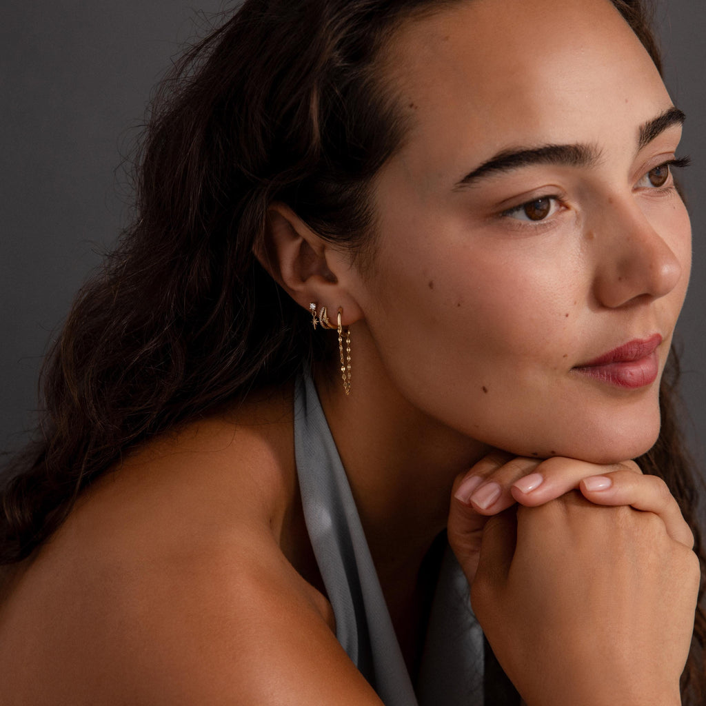 A woman with wavy hair, wearing a gray top and Mirror Chain Huggies, rests her chin on her hands and gazes to the side.