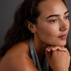 A woman with wavy hair, wearing a gray top and Mirror Chain Huggies, rests her chin on her hands and gazes to the side.