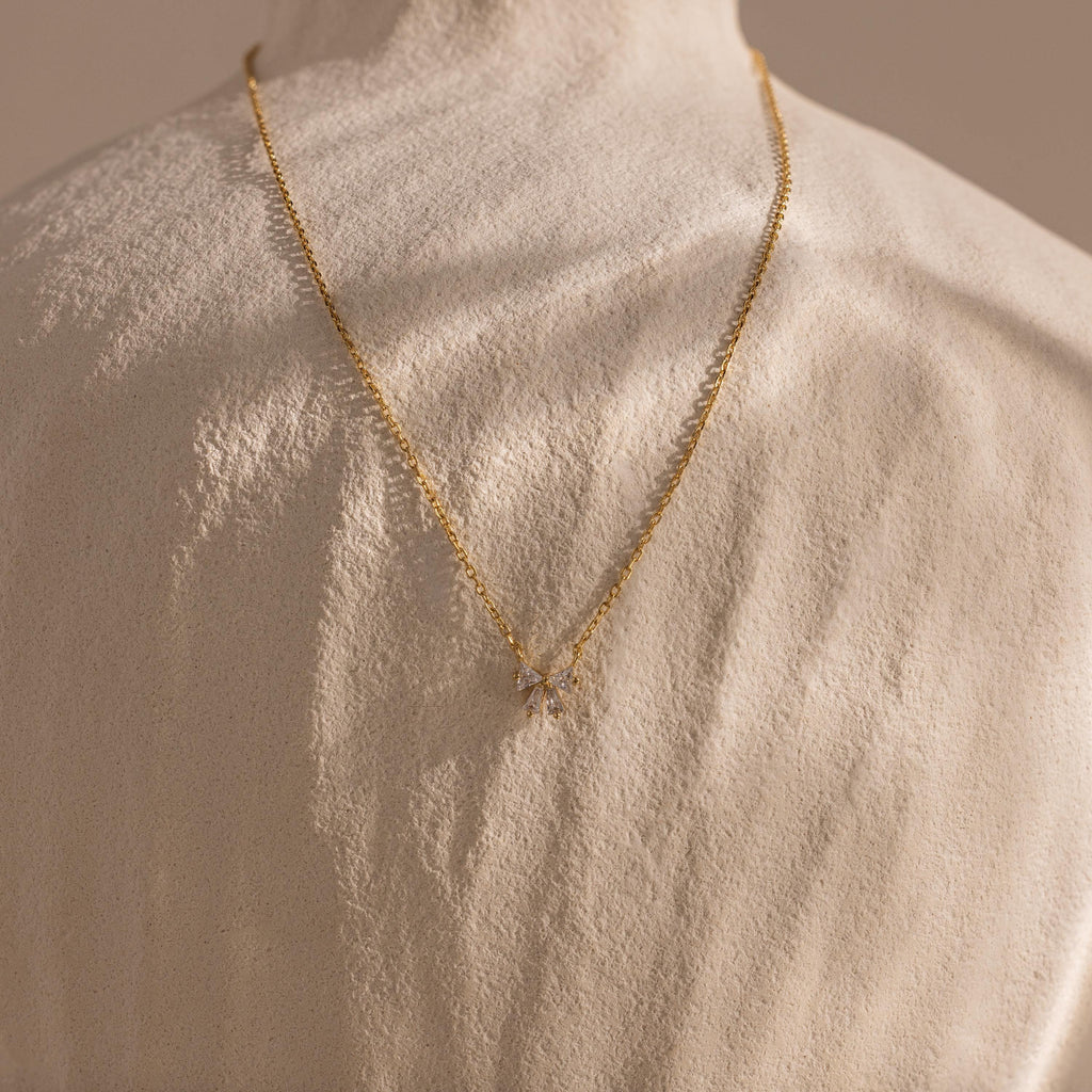 The Diamond Bow Necklace in 18K Gold, featuring a sparkling bow pendant, is draped over the back of a textured white sculpture.