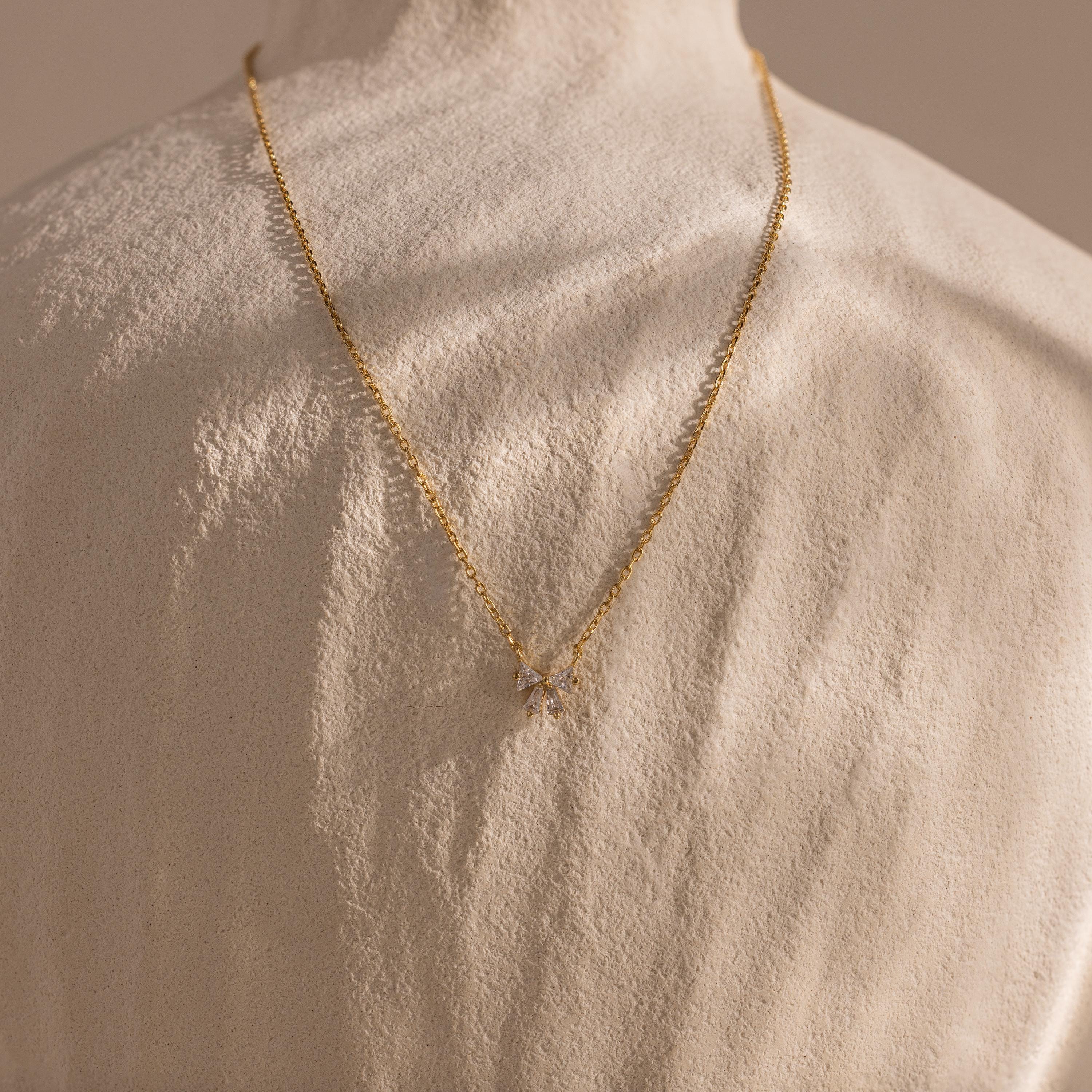 The Diamond Bow Necklace in 18K Gold, featuring a sparkling bow pendant, is draped over the back of a textured white sculpture.