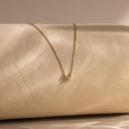 The Diamond Bow Necklace features a gold-plated, delicate bow pendant accented with sparkling cubic zirconia, displayed on a textured cream surface under soft lighting.