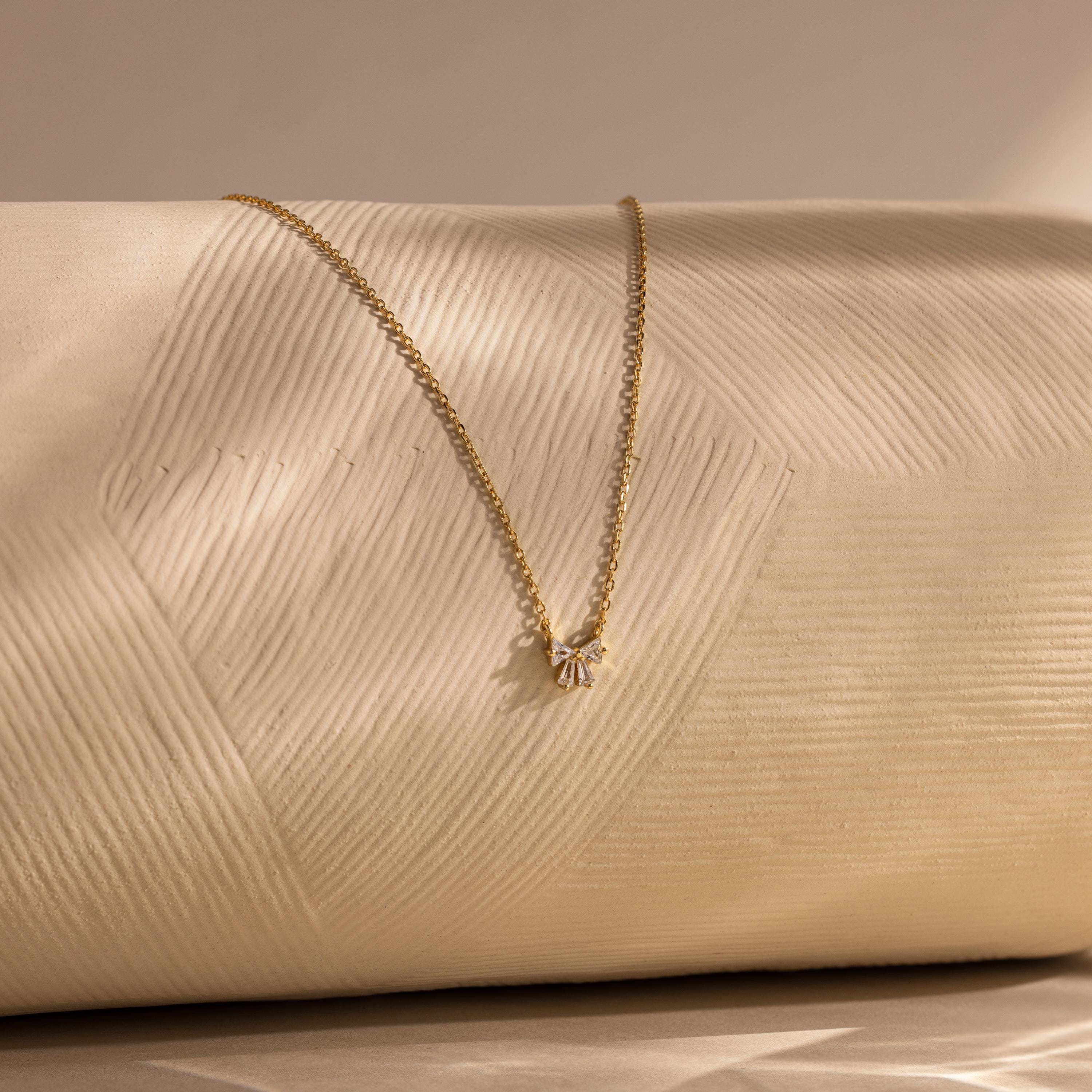 The Diamond Bow Necklace features a gold-plated, delicate bow pendant accented with sparkling cubic zirconia, displayed on a textured cream surface under soft lighting.