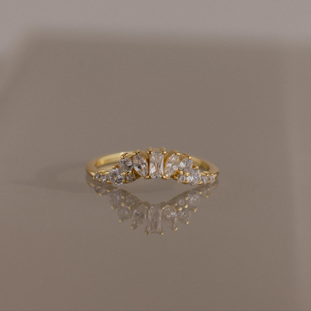 The Diamond Curve Cluster Ring in 18K Gold, adorned with a Baguette Stone in the center and a cluster of marquise and round diamonds on the sides, is shown on a reflective surface.