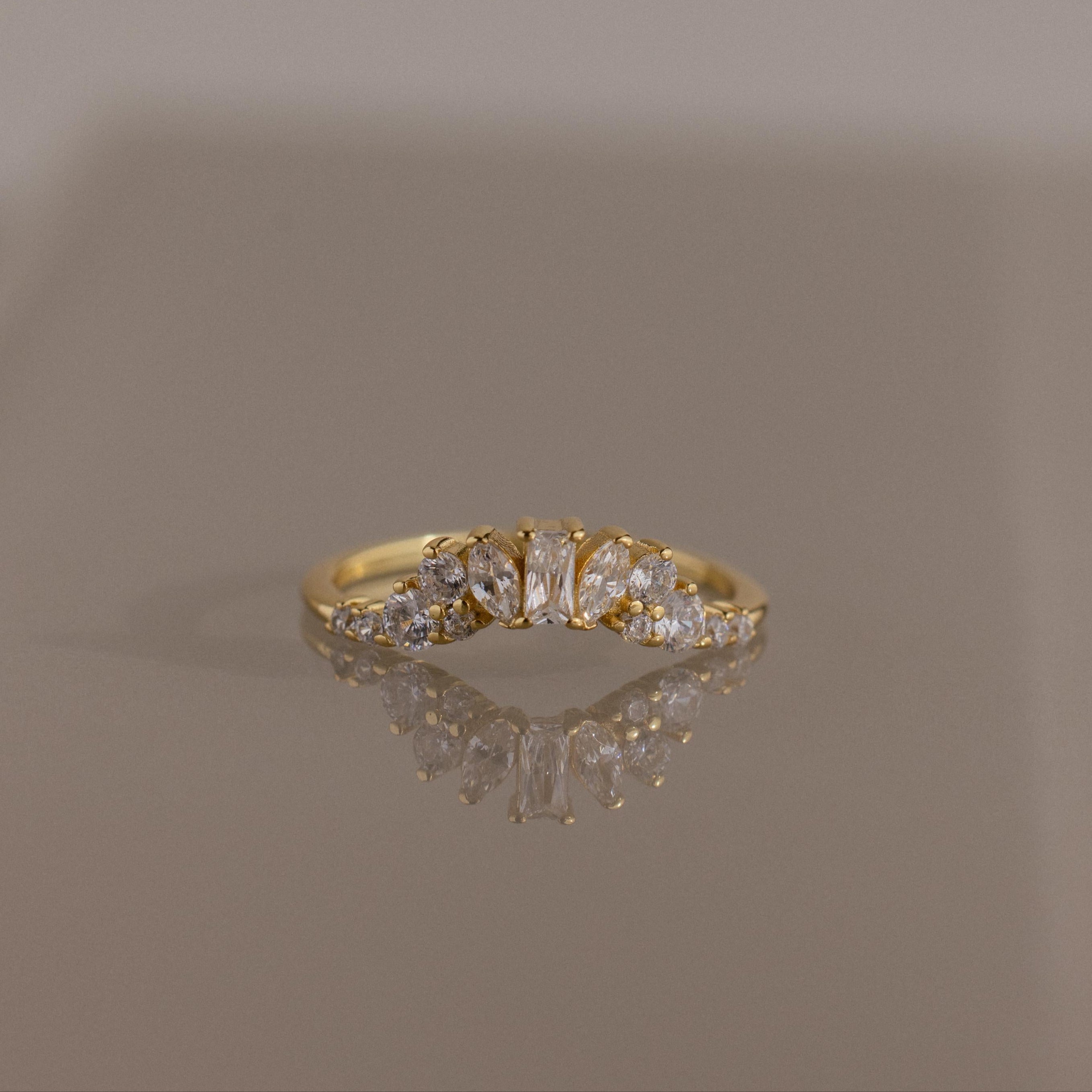 The Diamond Curve Cluster Ring in 18K Gold, adorned with a Baguette Stone in the center and a cluster of marquise and round diamonds on the sides, is shown on a reflective surface.