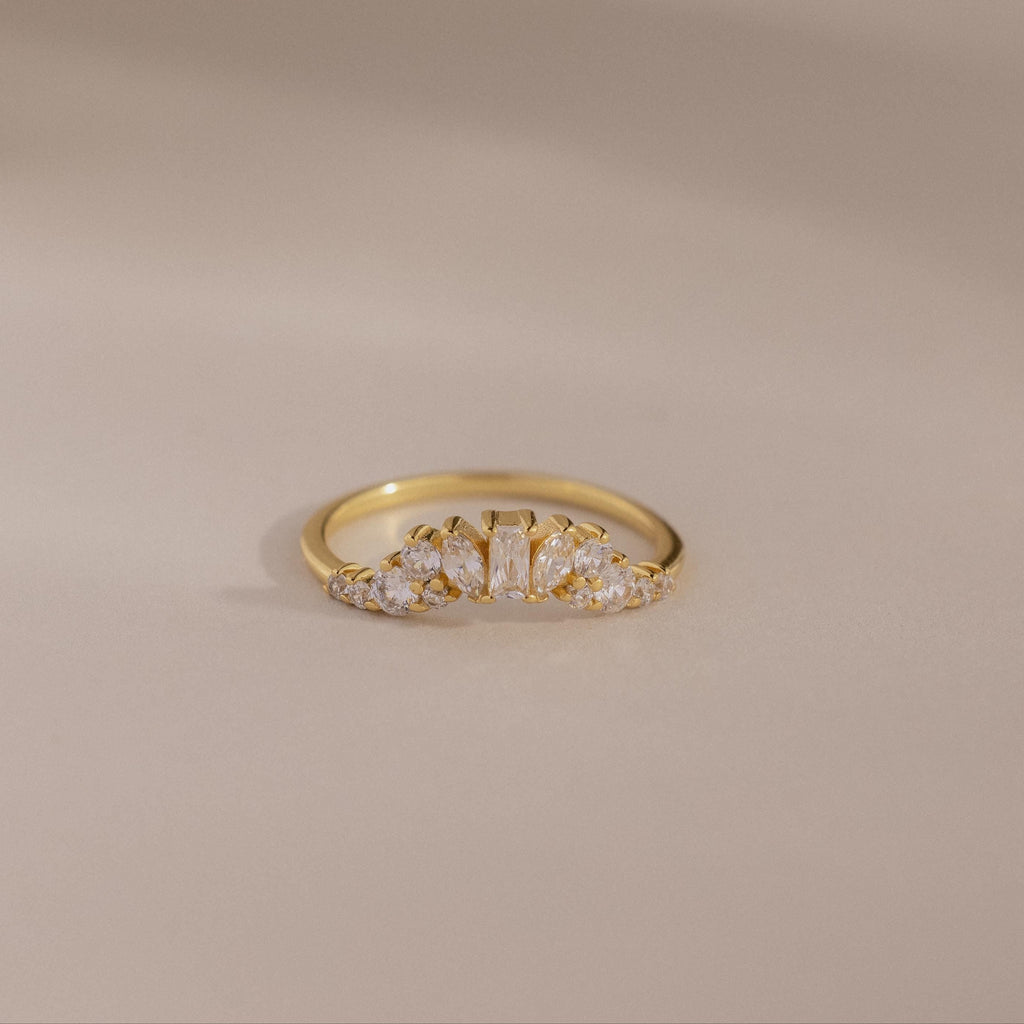 The Diamond Curve Cluster Ring features an baguette stone and a row of clear marquise and round diamonds set in gold, displayed on a smooth, neutral surface.