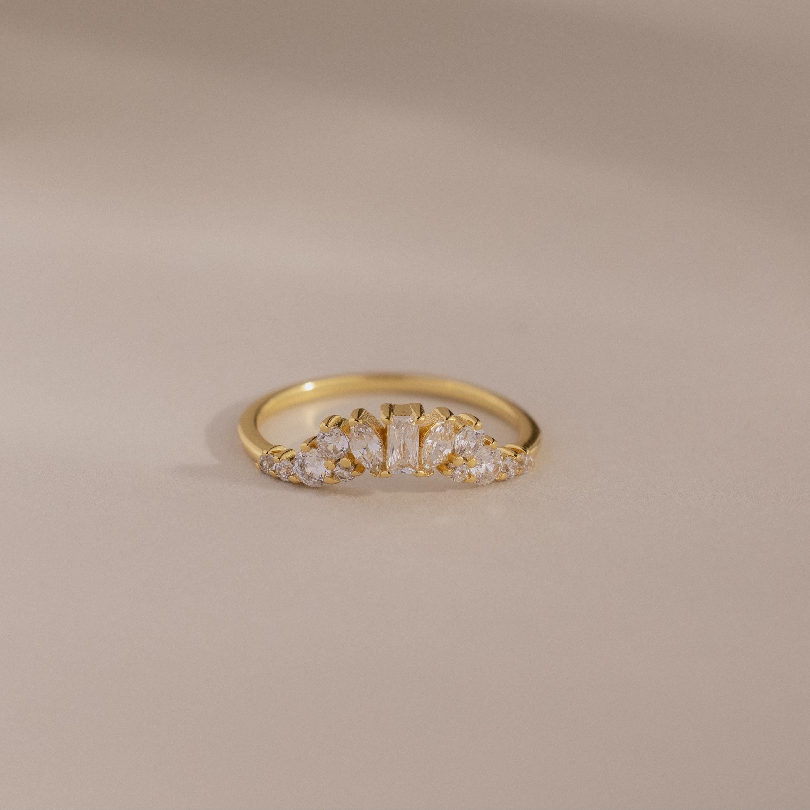 The Diamond Curve Cluster Ring features an baguette stone and a row of clear marquise and round diamonds set in gold, displayed on a smooth, neutral surface.