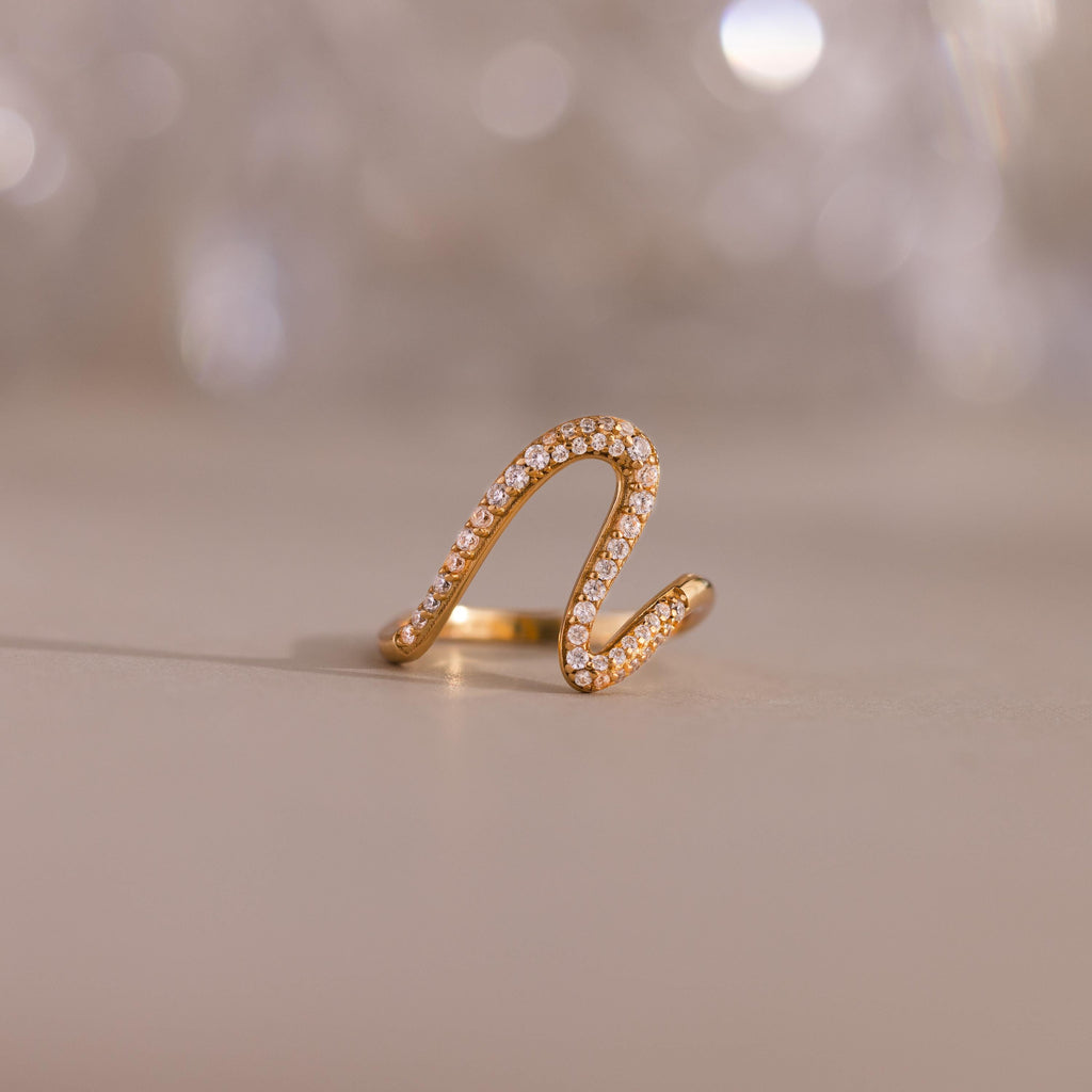 The Pave Wave Ring, crafted in gold and accented with pavé-set diamonds, is elegantly displayed against a smooth surface and blurred background. Ideal as a stacking ring for adding subtle sophistication to any look.