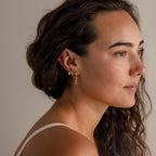 A woman with wavy brown hair wears Bow Drop Studs, delicate bow earrings, as she gazes to the side against a neutral background.