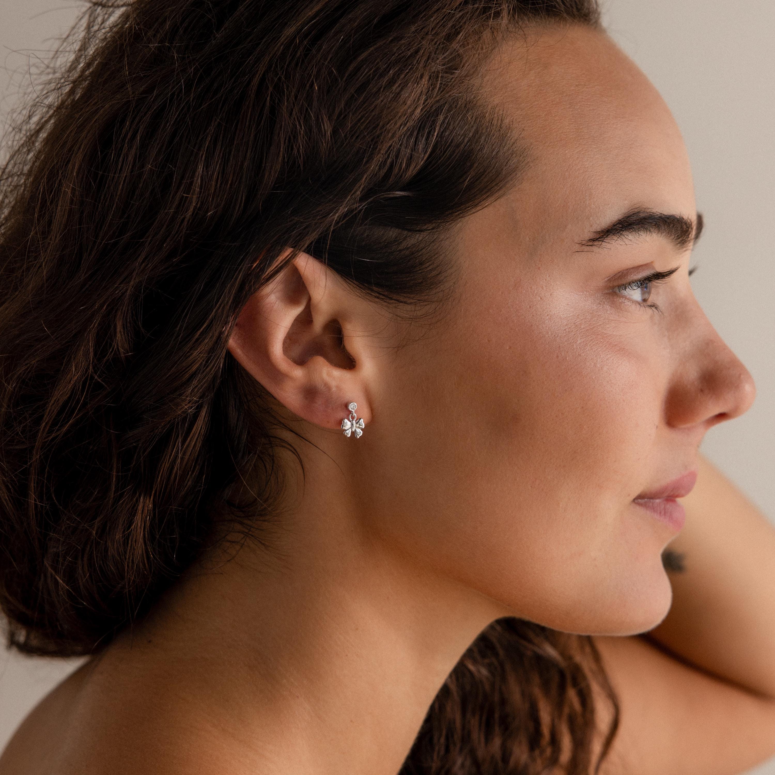 A woman with wavy brown hair wears Bow Drop Studs in Sterling Silver—delicate, floral-inspired earrings—while looking to the right.