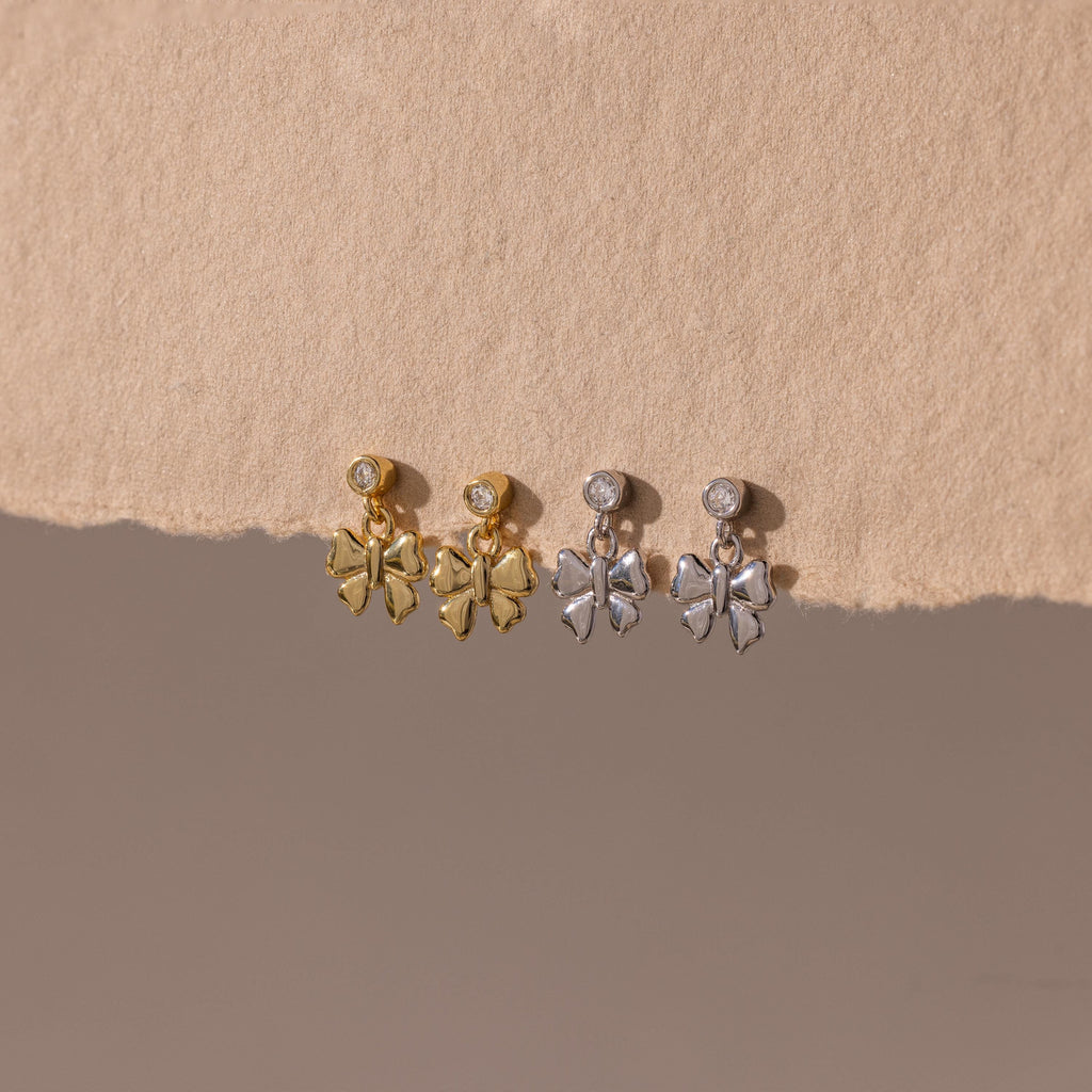 Two pairs of Bow Drop Studs—delicate dangling earrings in gold and silver—are displayed on a beige textured surface.