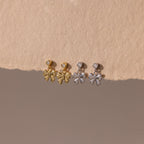 Two pairs of Bow Drop Studs—delicate dangling earrings in gold and silver—are displayed on a beige textured surface.