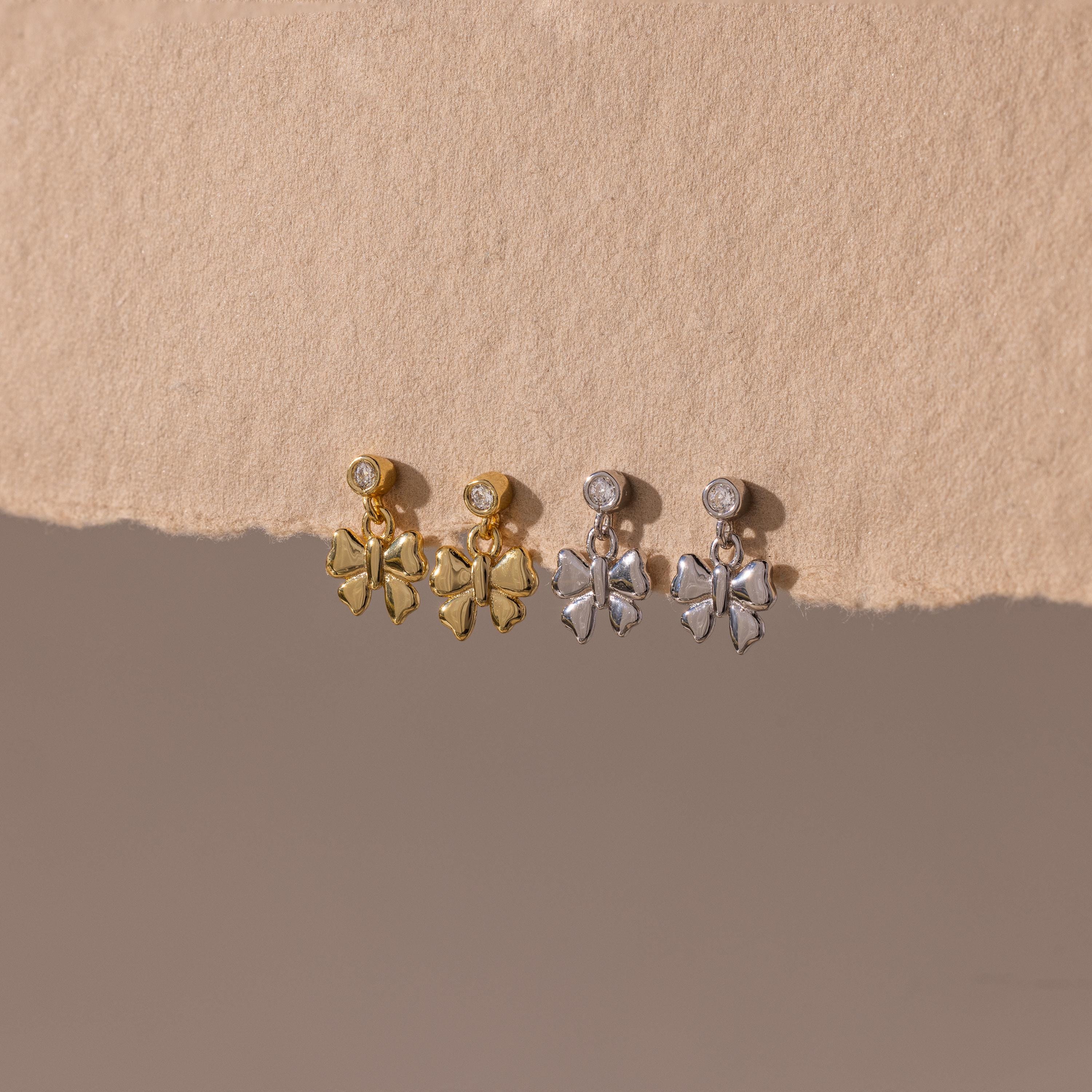 Two pairs of Bow Drop Studs—delicate dangling earrings in gold and silver—are displayed on a beige textured surface.