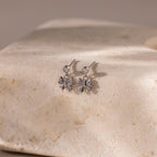 Bow Drop Studs in Sterling Silver, featuring a delicate bow design with small clear stones, perfect for lovers of subtle jewelry, displayed on a textured beige stone surface.