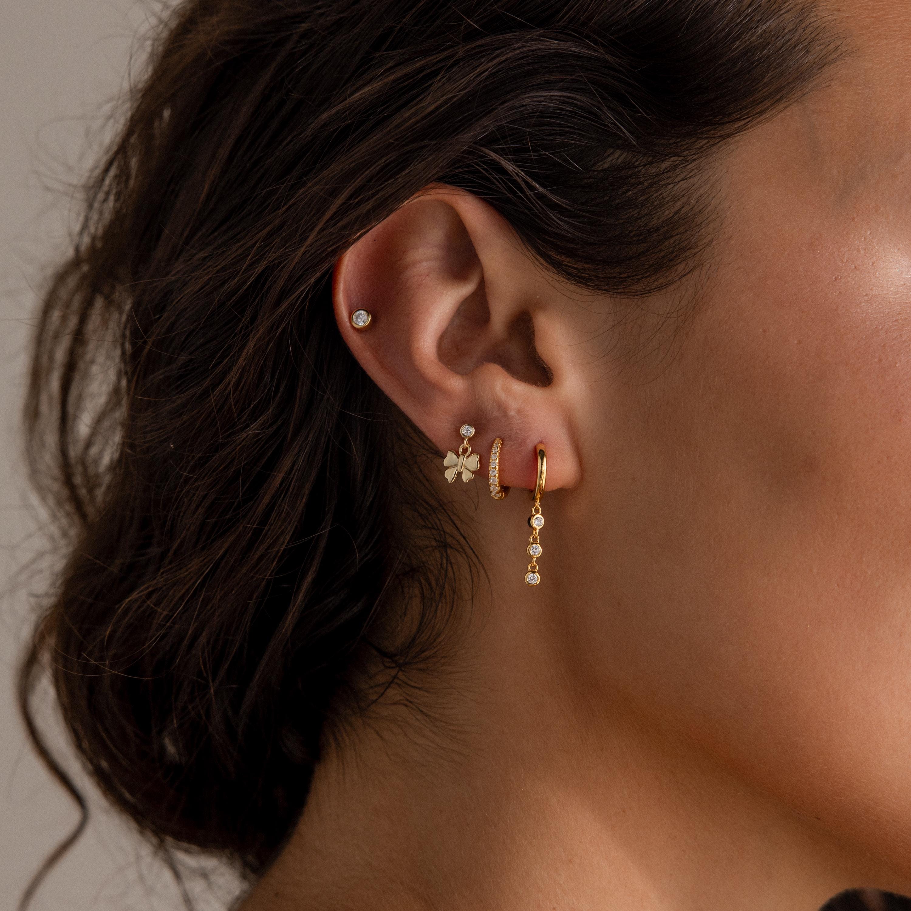 Close-up of a womans ear styled with multiple gold earrings, including the Bow Drop Studs in 18K Gold, alongside a stud, flower, and hoop for a chic, charming look.