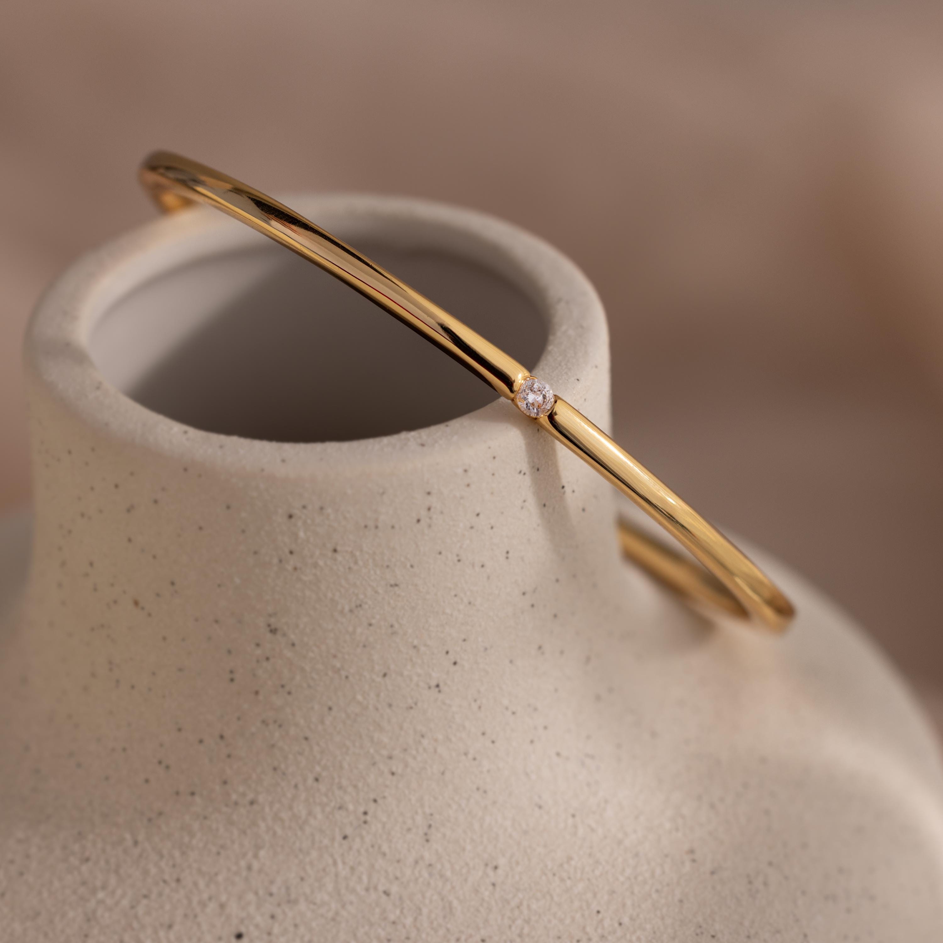 A Skinny Diamond Bangle in 18K Gold with a delicate diamond detail rests gracefully on a smooth, speckled ceramic vase.