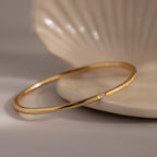 The Skinny Diamond Bangle—a minimalist bracelet with a thin gold band and single diamond—rests elegantly on a light-colored shell dish.