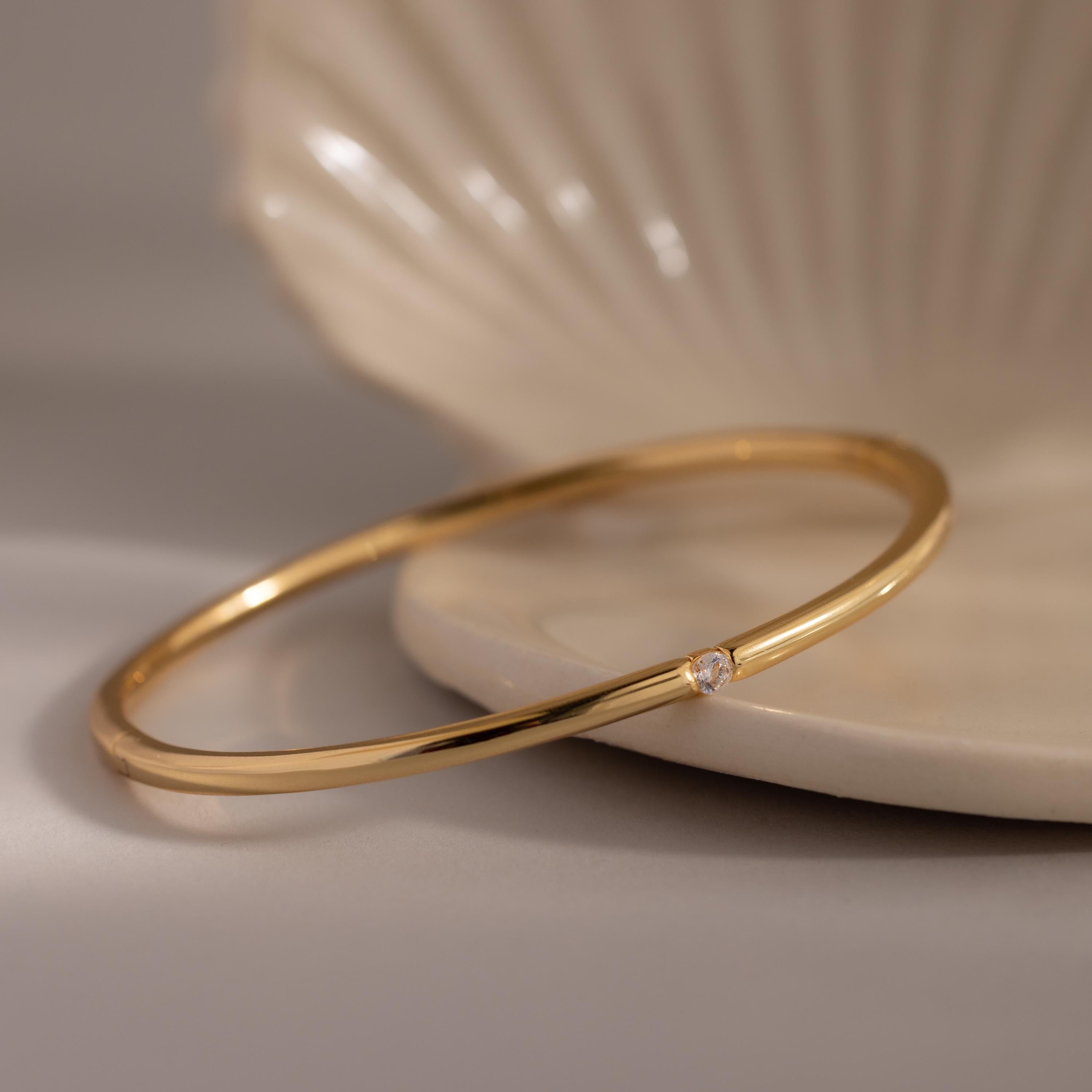 The Skinny Diamond Bangle—a minimalist bracelet with a thin gold band and single diamond—rests elegantly on a light-colored shell dish.