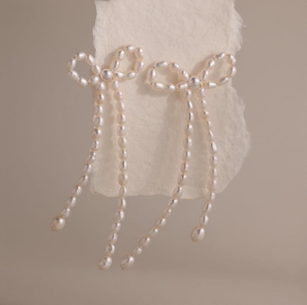 The Pearl Bow Earrings, shaped like elegant bows and adorned with pearls, are displayed on textured white paper against a neutral background, adding a sophisticated touch to any outfit.