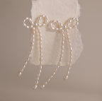 The Pearl Bow Earrings, shaped like elegant bows and adorned with pearls, are displayed on textured white paper against a neutral background, adding a sophisticated touch to any outfit.