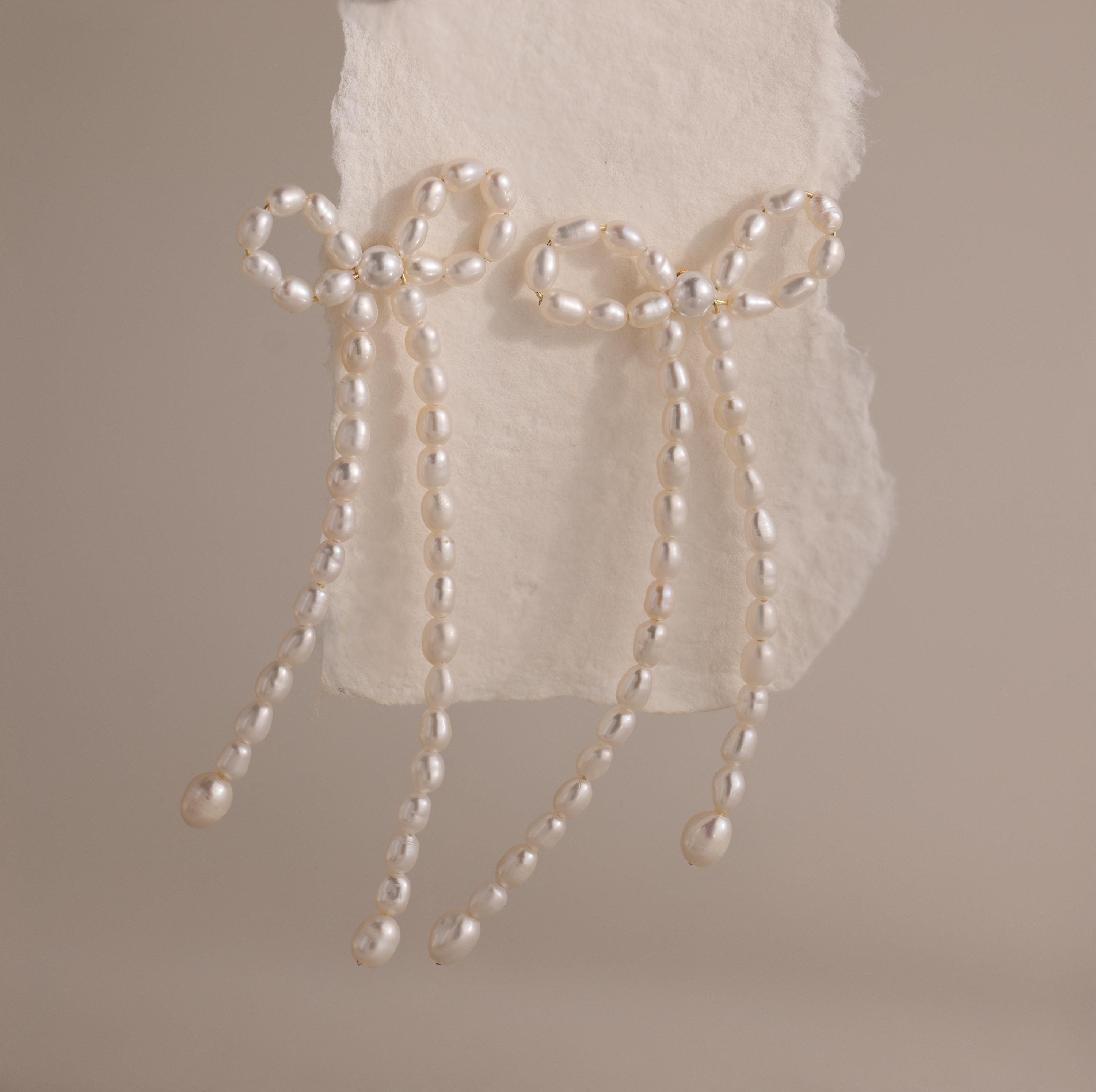 The Pearl Bow Earrings, shaped like elegant bows and adorned with pearls, are displayed on textured white paper against a neutral background, adding a sophisticated touch to any outfit.