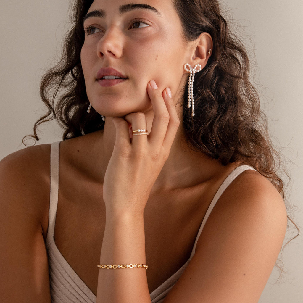 A woman with long curly hair wears Pearl Bow Earrings, a gold bracelet, and a ring as she gazes thoughtfully—showcasing elegant jewelry and timeless style.